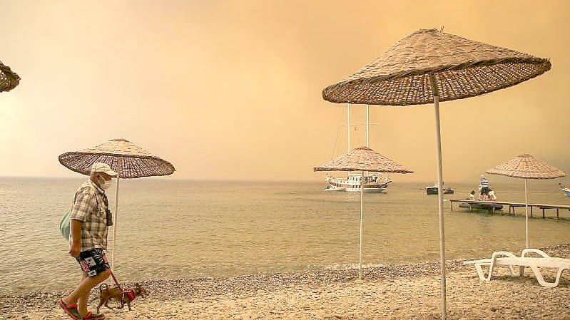 Ein Mann spaziert mit seinem Hund durch rauchverhangene Luft an einem türkischen Strand. Foto: Emre Tazegul/AP/dpa