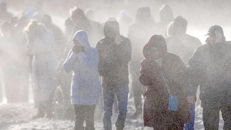 Schaulustige gehen durch den Sandsturm auf die Mole von Warnemünde. Urlauber und Tagesgäste beobachten bei Sonnenschein und kräftigem Wind den Wellengang durch das Sturmtief „Nadia“. Foto: Jens Büttner/dpa-Zentralbild/dpa