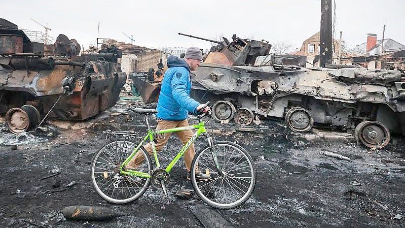 Ein Mann in Butscha schiebt sein Fahrrad an den Überresten eines russischen Militärfahrzeugs vorbei. Foto: Serhii Nuzhnenko/AP/dpa