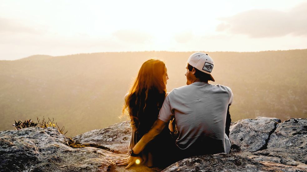 Ein junges Pärchen sitzt zusammen auf einem Berg und schaut sich den Sonnenuntergang an. Foto: Unsplash/Cody Black